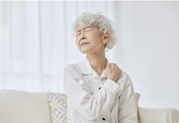 Senior woman experiencing shoulder pain and muscle tension while sitting on a sofa.