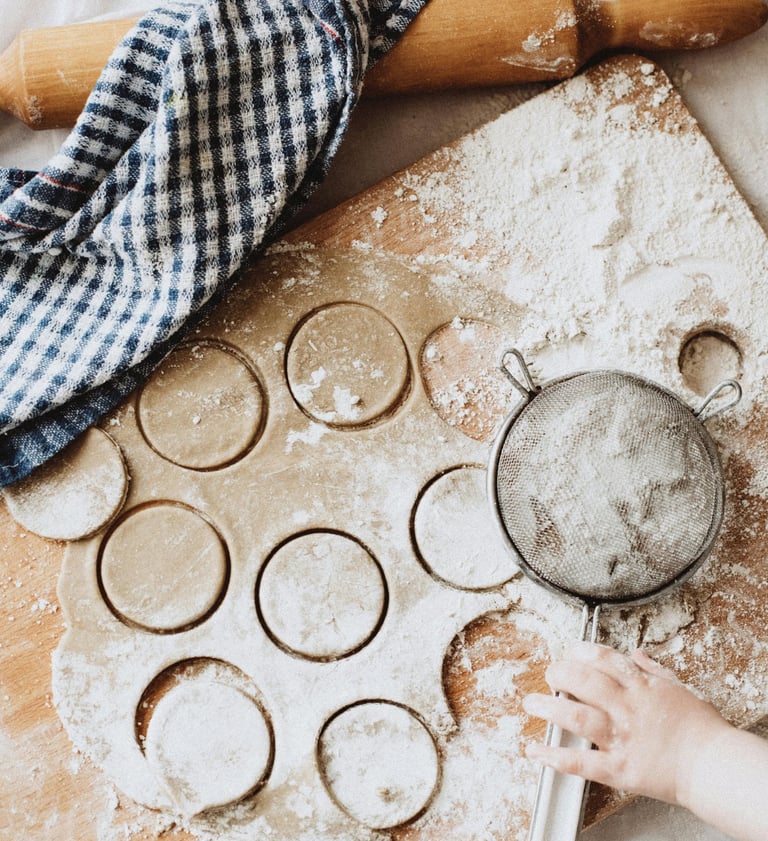 A child's hand using a flour sifter on cookie dough cut into circles on a wooden baking board.