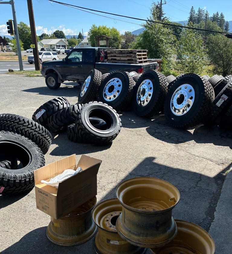 A collection of heavy-duty truck tires and chrome rims displayed on asphalt at a tire service shop.