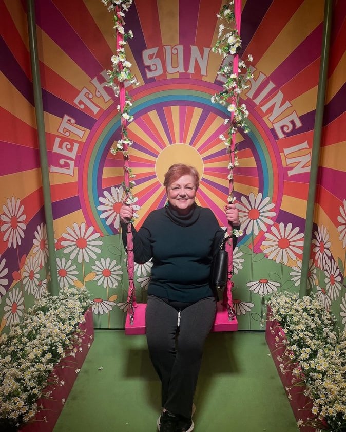 Photo of Debbie, a white woman with short red & blonde hair, on a flowery swing in front of a sign: "Let the Sunshine In."