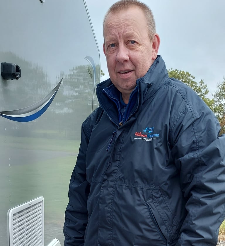 William Wilson in a Wilson Caravan Towing jacket standing next to a silver caravan outdoors.