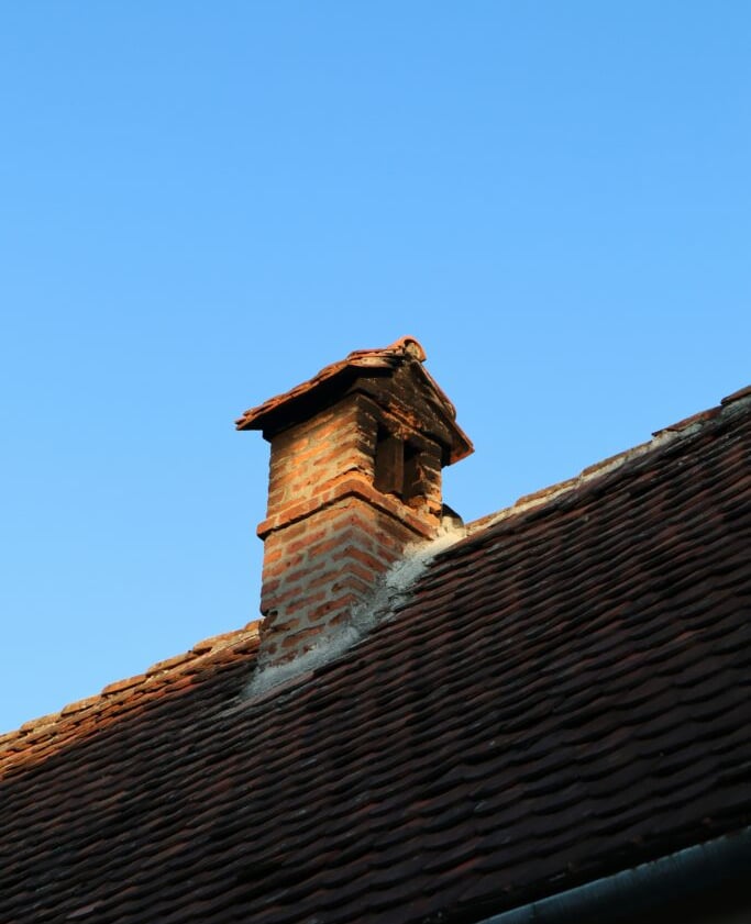  A brick chimney with a cap on a tiled roof against a clear blue sky, suitable for chimney caps, cro