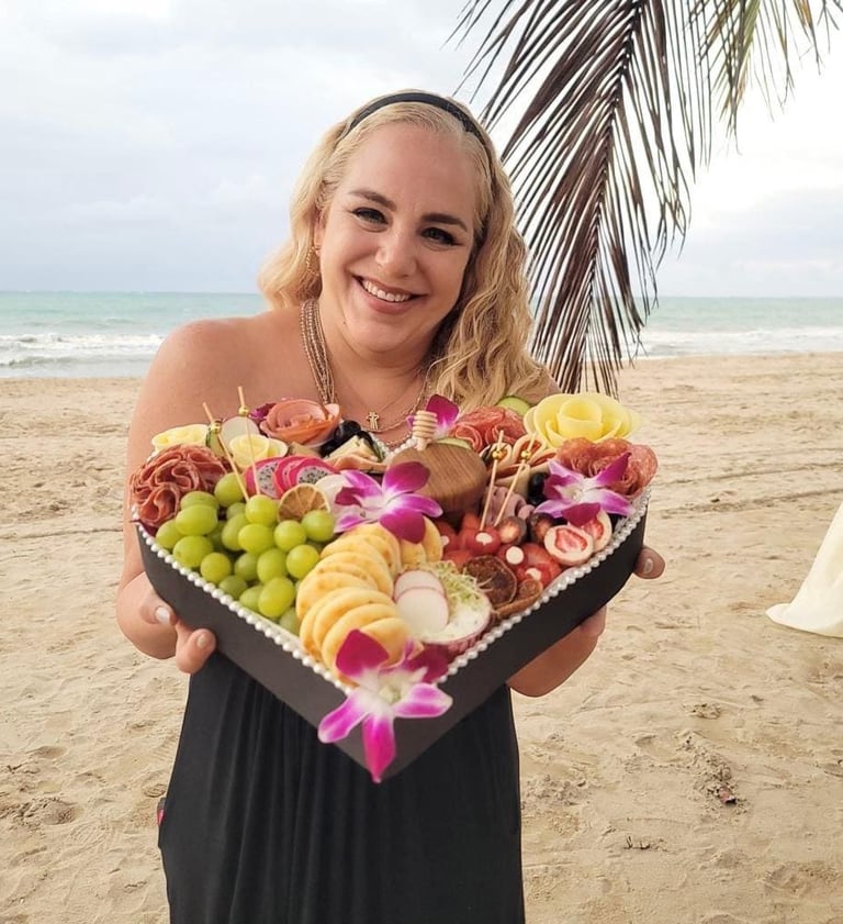 Una mujer sonriente en una playa tropical sosteniendo una tabla de charcutería en forma de corazón.