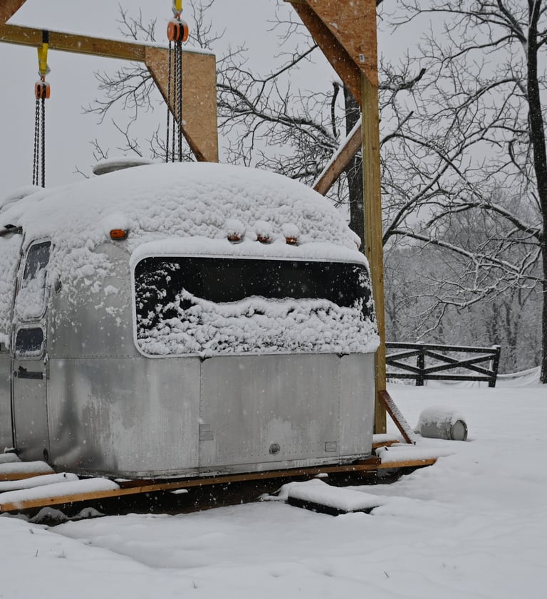 Snowcovered shell of the airstream sitting on the lumber. 