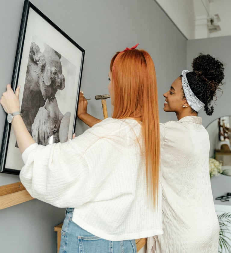 Two women installing framed refugee artwork in a gallery space.