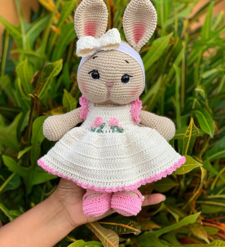 Handmade amigurumi bunny doll wearing a white crochet dress and pink shoes outdoors.