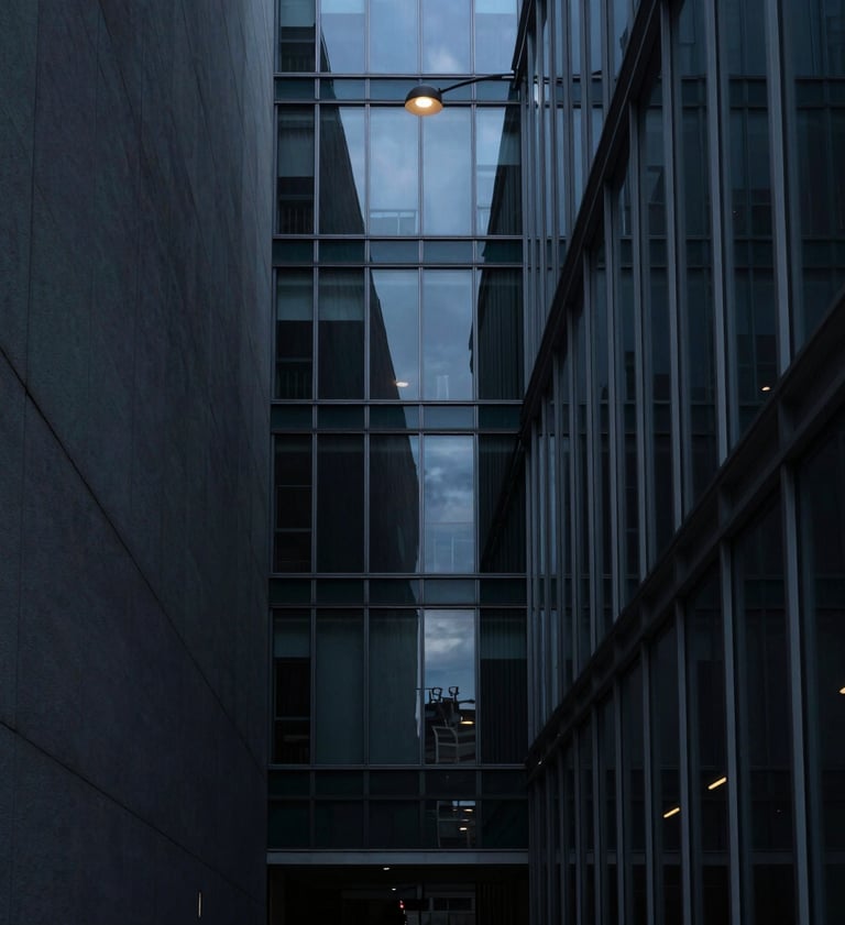 A minimalist vertical photography shot of a narrow alleyway between modern glass buildings. The palette features deep slate and muted blue tones. A single overhead lamp provides a pool of soft light against the dark shadows. International / Western metropolitan setting.