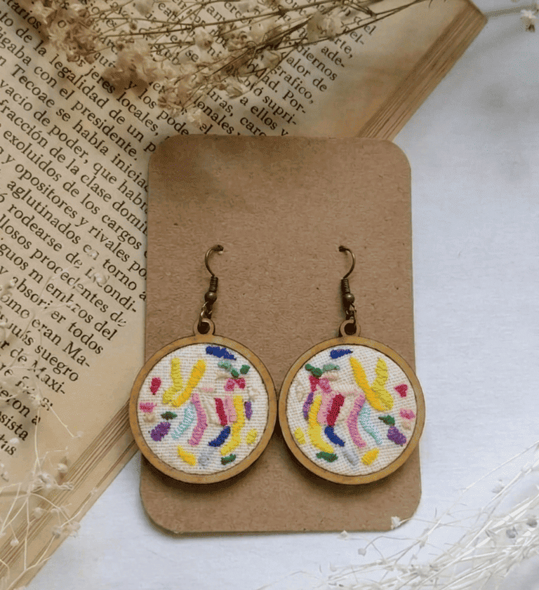 Pendientes de aro con bordado Tenango hecho a mano, con hilos coloridos sobre un fondo vintage.