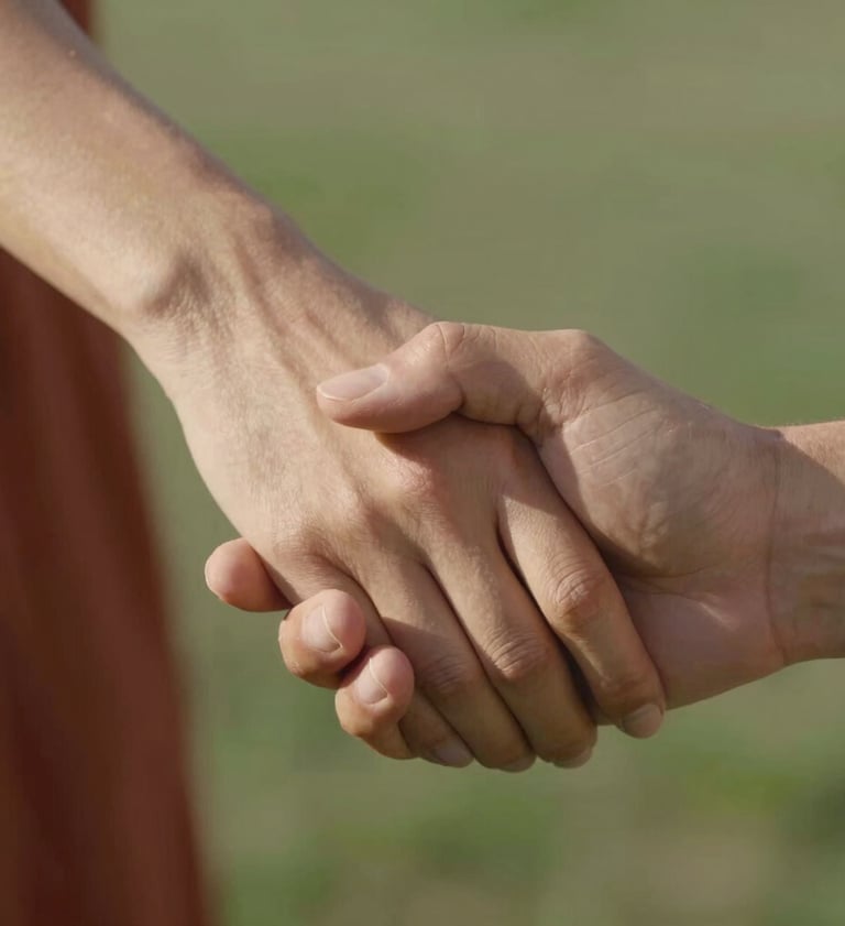 A close-up photograph of a couple's hands gently intertwined, focusing on the texture of their skin and the soft glow of a North American afternoon sun. The background is a soft blur of terracotta and sage green colors, emphasizing the intimacy of the touch.