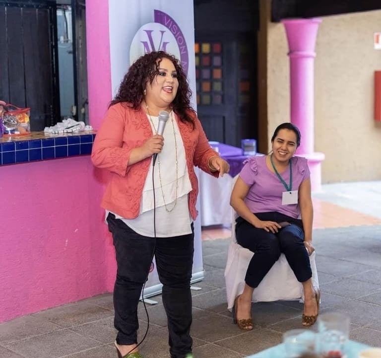 Una oradora sonriente con cabello rizado sosteniendo un micrófono y dando una presentación en vivo