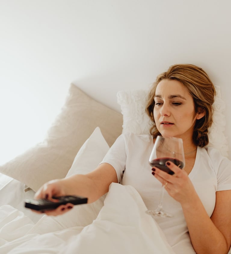 A woman relaxes in bed holding a glass of red wine and using a TV remote.