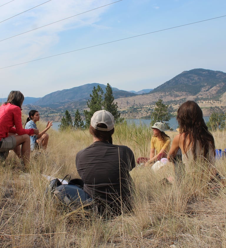 A group of people sitting on a hill top with Skaha Lake and rolling mountains in the background.