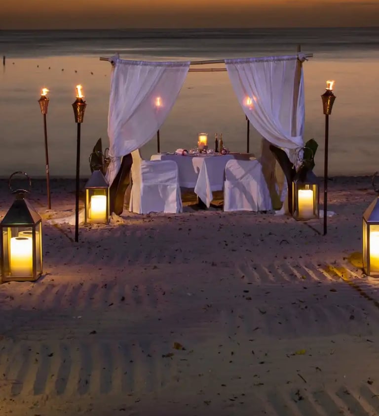 Romantic private dinner for two on a sandy beach at sunset with lanterns and tiki torches.