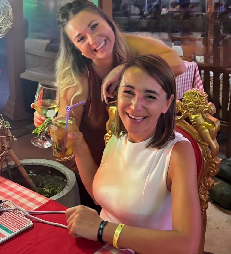 Two smiling women enjoying cocktails and white wine at an outdoor restaurant dinner table.