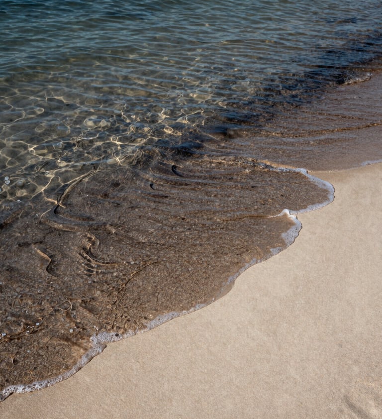 A candid shot of the ocean tide just beginning to wash over a sand pattern. The water is clear, showing the transition between the charcoal-colored wet sand and the light cream-colored dry soft sand.