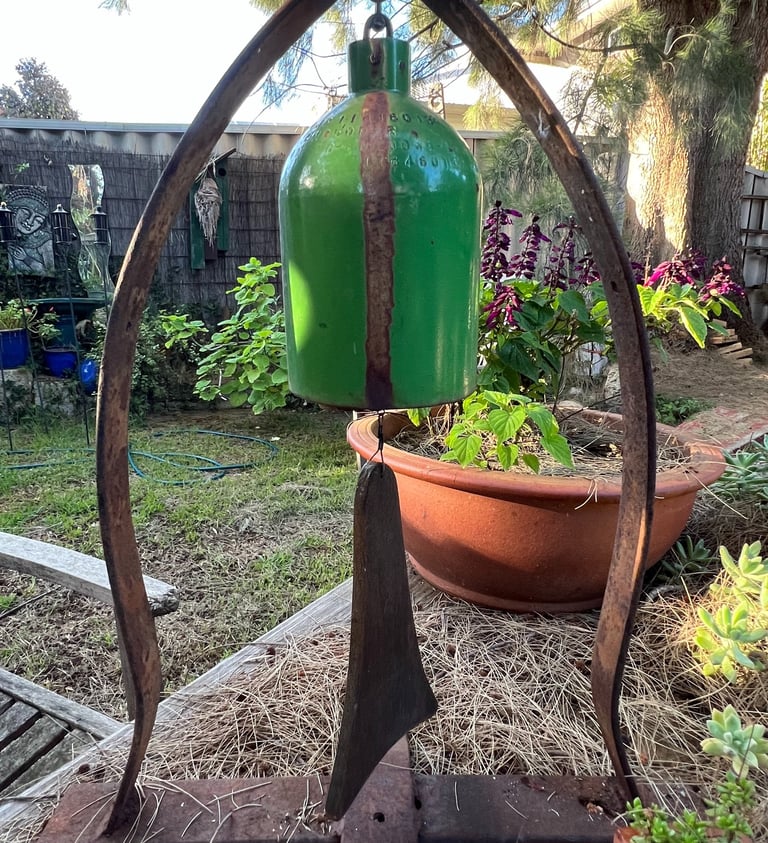 Rustic green metal garden bell wind chime hanging on a weathered iron stand in a backyard garden.