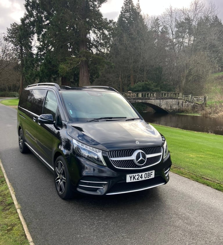 Black Mercedes-Benz V-Class luxury MPV parked on a scenic road near a stone bridge and river.