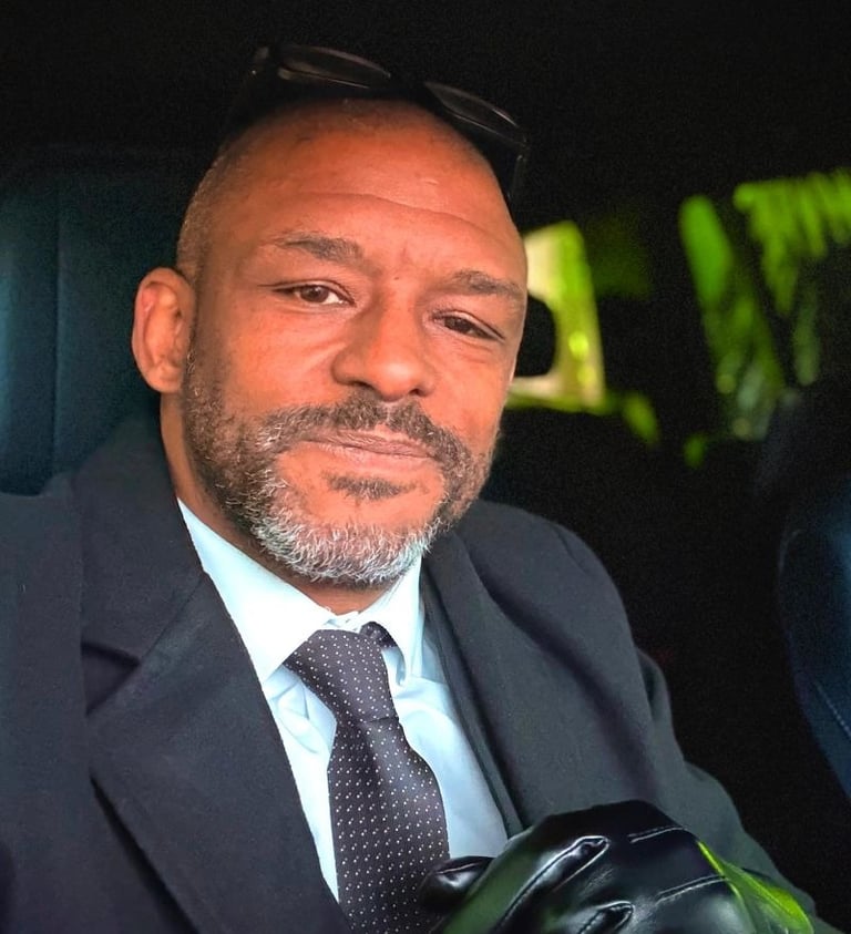 Bradley in a black suit, tie, and leather gloves sitting inside a car.
