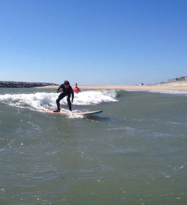 A young male surfer rides an unbroken wave with sunny skies while a surf instructor looks on