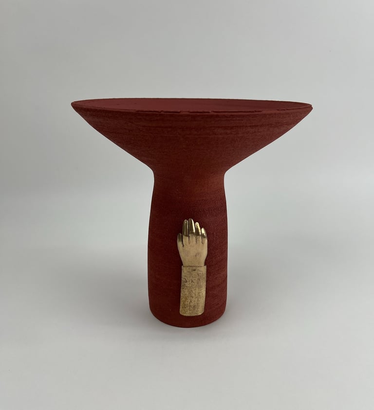 A tall thrown red ceramic vessel, wide at the top. A cast bronze hand is fixed on its base.
