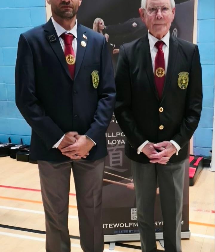 Two karate officials in blazers and ties standing in front of a White Wolf martial arts banner.