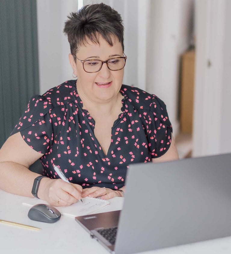 Professional woman with glasses working on a laptop and taking notes in a home office.