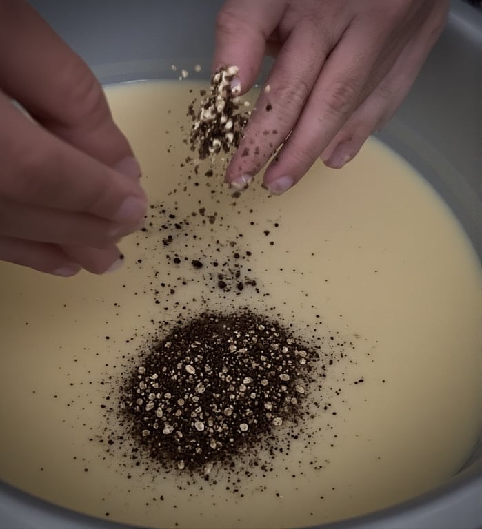 Hands sprinkling coffee grounds and oats into melted soap mixture