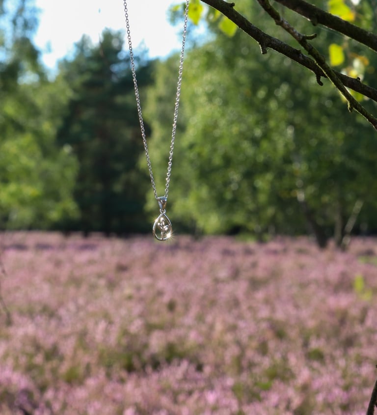 HeideSchmuck Silberkette mit tropfenförmigem Kristallanhänger, aufgehängt an einem Ast vor blühendem Heidekraut in der Natur.