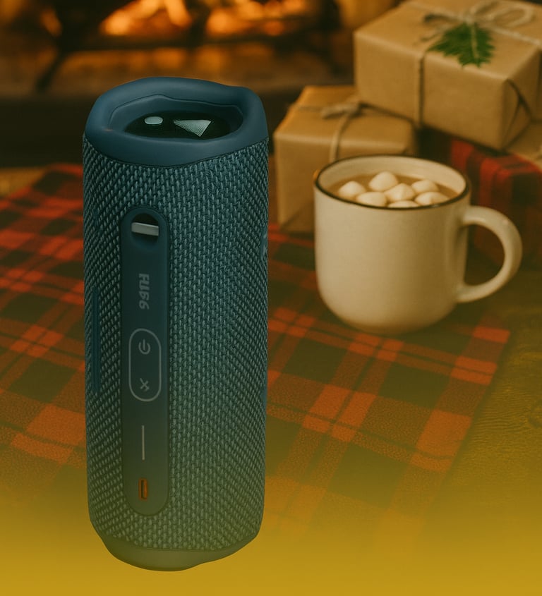 JBL Flip 6 Bluetooth speaker on plaid blanket with hot cocoa and wrapped gifts by a fireplace.
