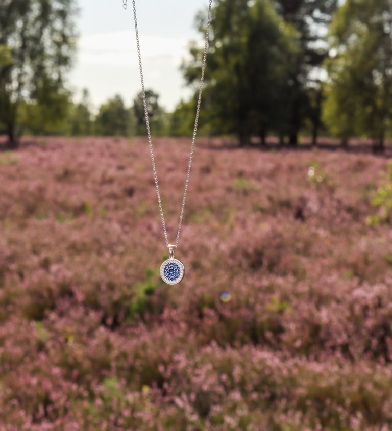 HeideSchmuck Silberkette mit rundem blauen Medaillon, gehalten vor blühender Heide in der Natur.