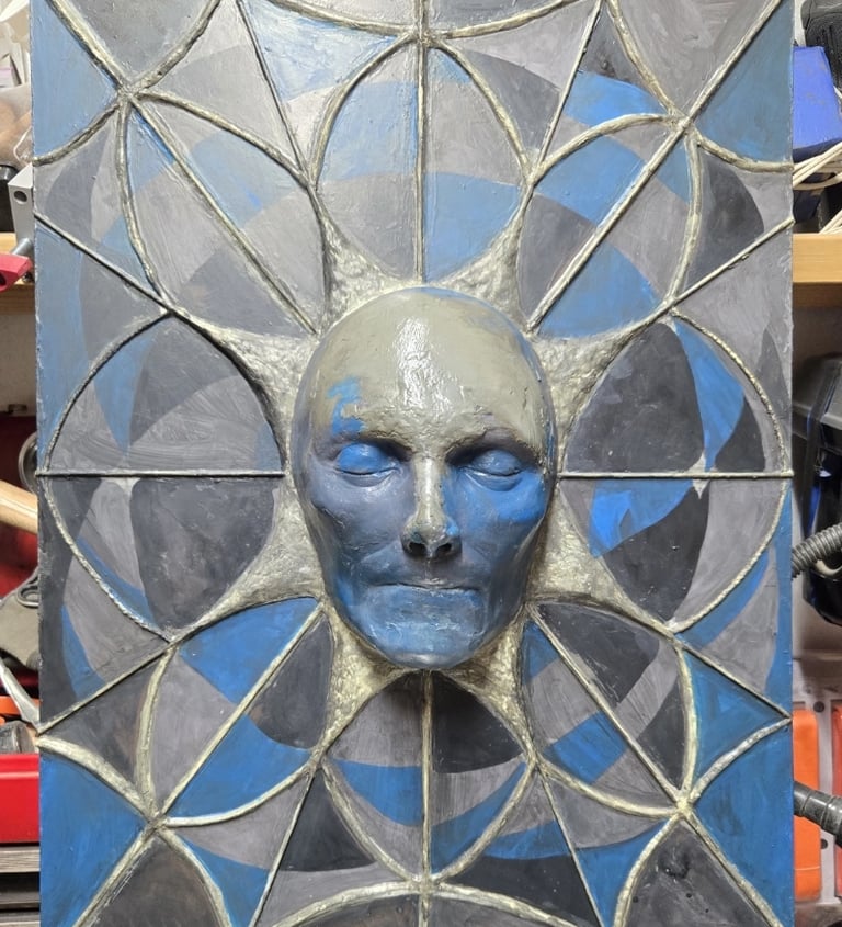 wall hanging relief sculpture in earthy greens, blues and blacks. A face protruding from a geometric web design.