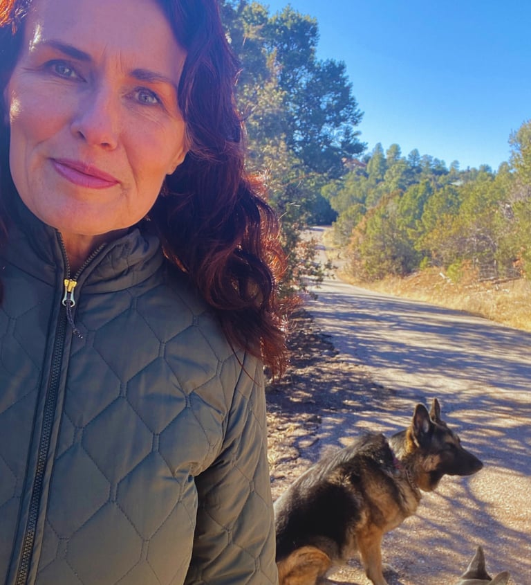 A woman in a quilted jacket hiking with her German Shepherd on a sunny trail.