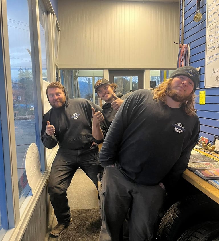 Three cheerful auto shop mechanics in black hoodies posing and giving thumbs up in a garage office.