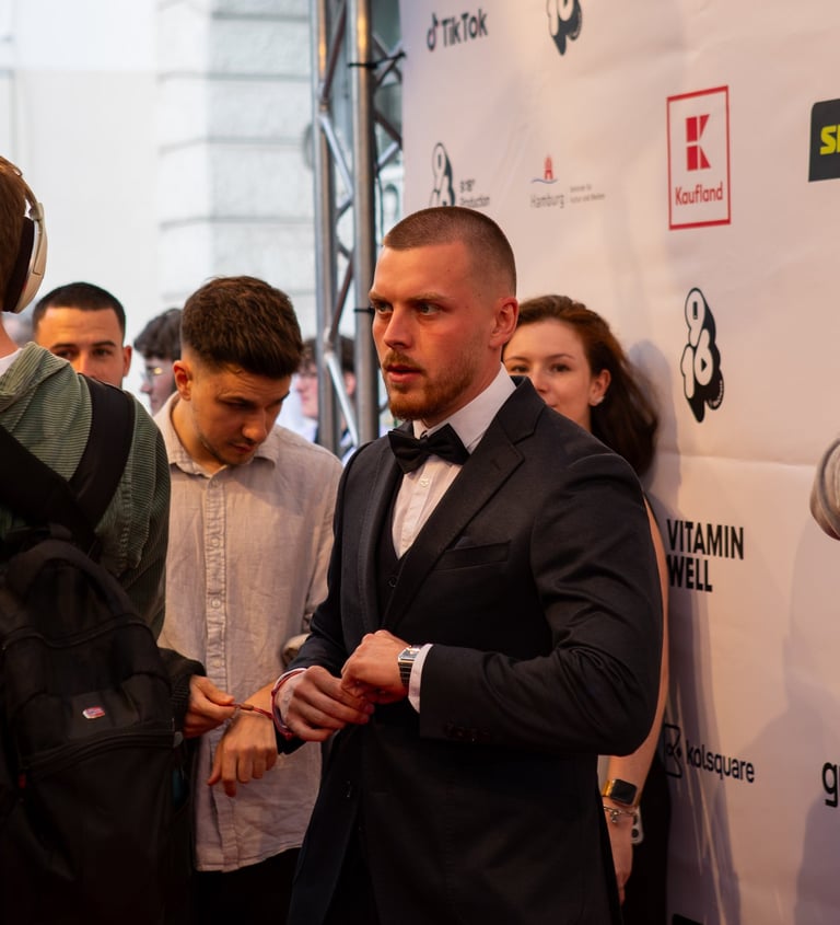 A man in a navy suit and bowtie poses on a red carpet in front of a brand sponsor backdrop.