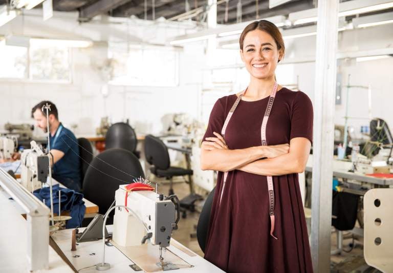Diseñadora de moda sonriente de pie en una fábrica de confección con máquinas de coser industriales