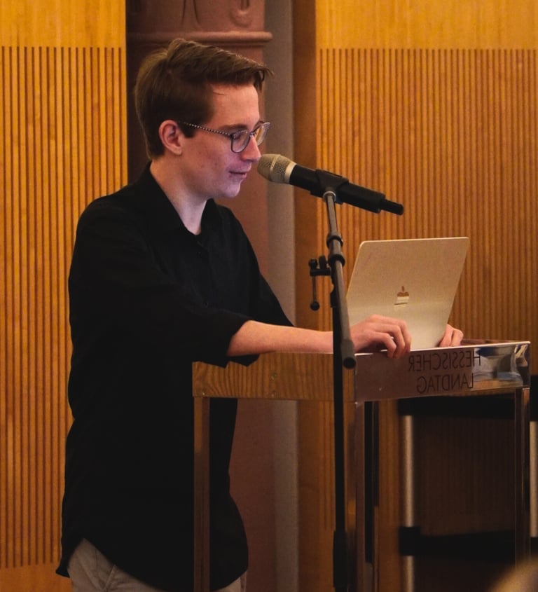 Junger Mann mit Brille hält Vortrag am Laptop und spricht in Mikrofon auf Bühne – Rede