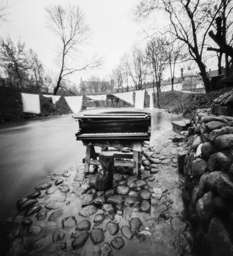 A pinhole captures a black and white photo of a piano gracefully placed on the side of a serene river