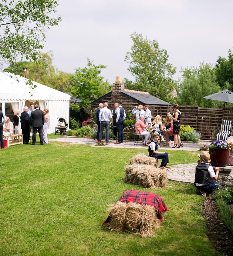Guests mingling at an outdoor garden wedding reception with a white marquee tent and hay bale seating.