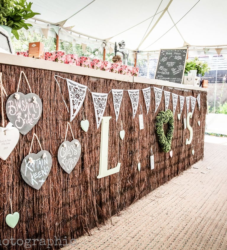 Rustic outdoor wedding bar with heart-shaped signs and white bunting under a marquee tent.