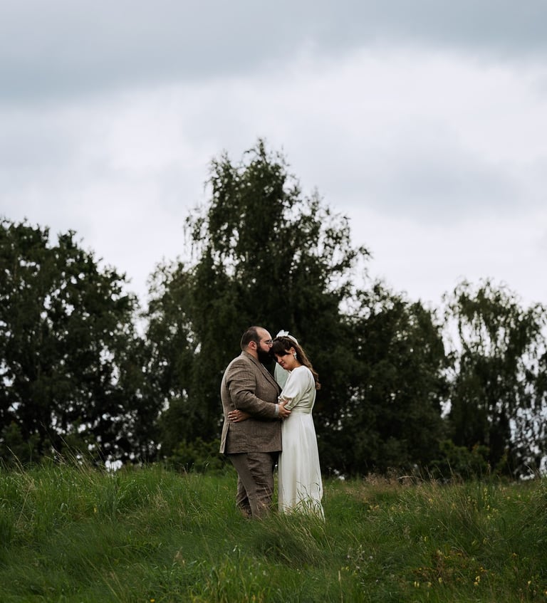 Bröllopspar som håller om varandra i högt gräs på en äng med skogen bakom.