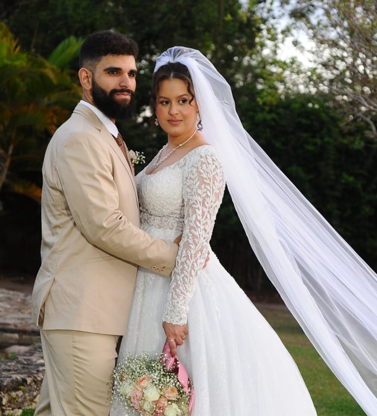 Uma noiva em um vestido de renda e véu longo posando ao ar livre com seu noivo de terno bege.