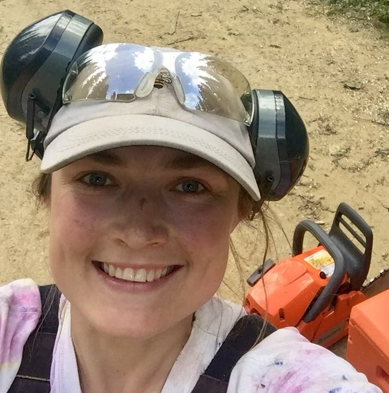 Keely smiles wide, wearing a beige ballcap and headphones. Behind her is an orange chainsaw.