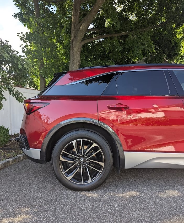 Side profile of a red Acura MDX luxury SUV parked on a paved driveway under a leafy green tree.