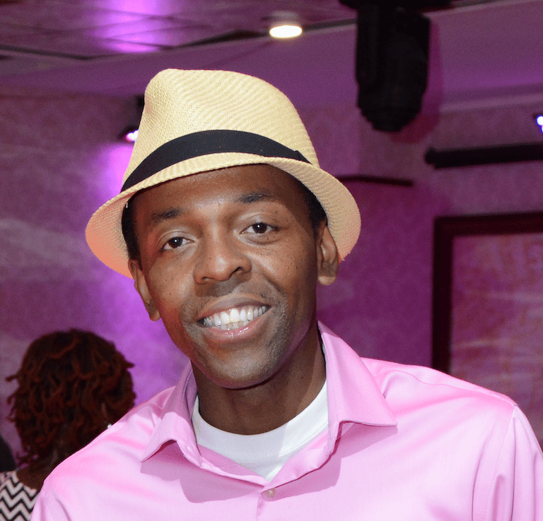 Zangba Thomson smiling in a pink button-down shirt and straw fedora hat at an indoor event.