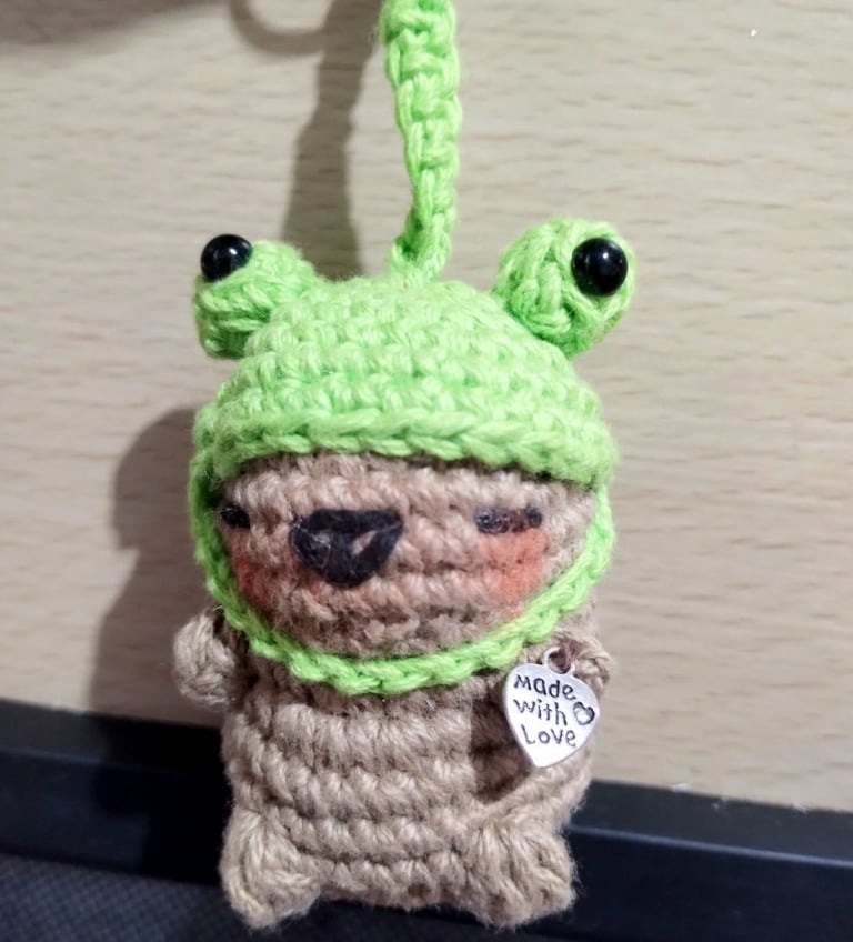 Llavero de capibara tejido a crochet, con un gorro de rana verde y un dije de corazón plateado.