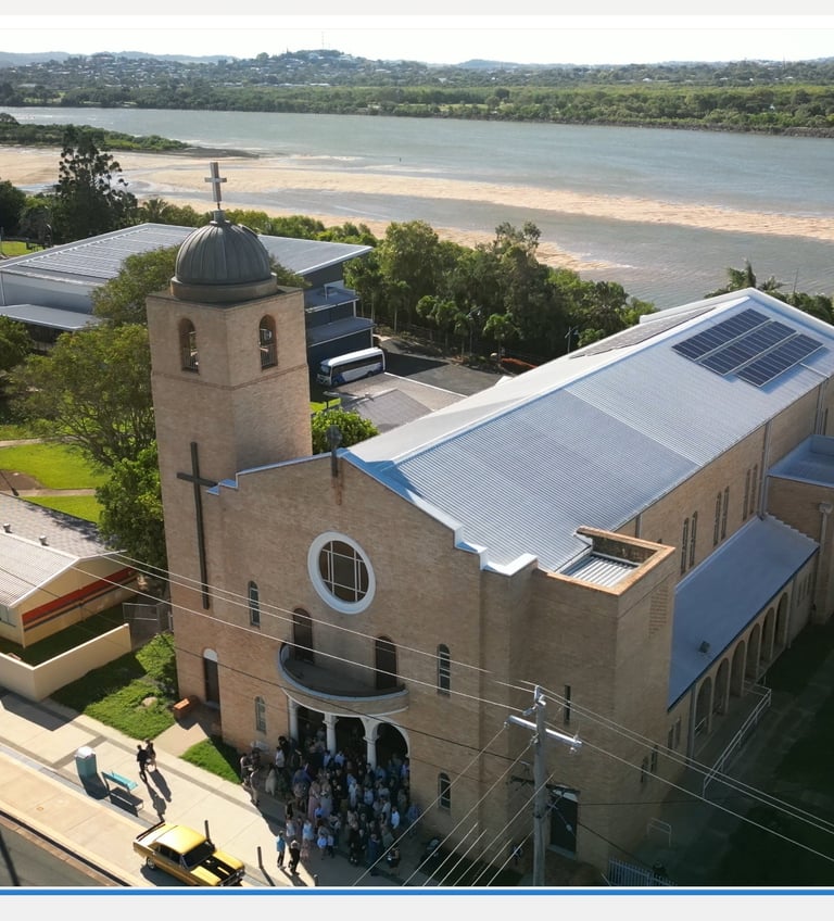 Aerial view of a brick church with solar panels on the roof near a river in a coastal town.