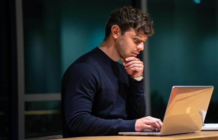 Individual seated at table with hand on chin, concentrating on a laptop in a dimly lit room.