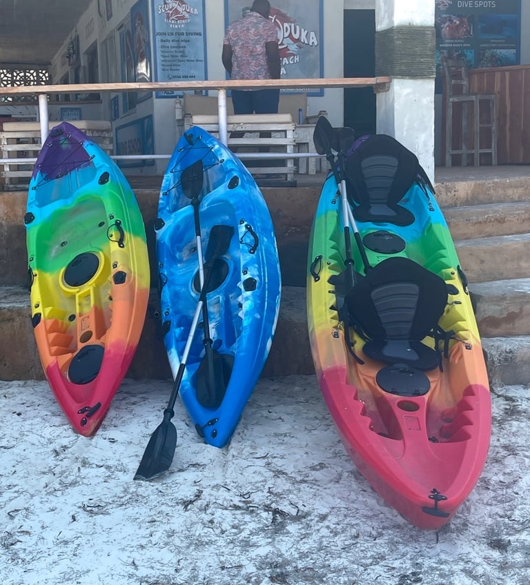 Three colorful sit-on-top kayaks leaning against a PADI diving school wall on a sandy beach.