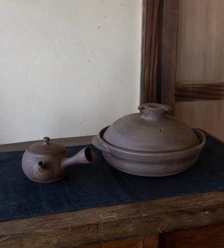 Handmade brown ceramic Japanese kyusu teapot and donabe clay pot on a rustic wooden table.