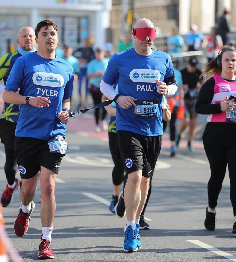 Two runners side by side, one is blindfolded, both are holding a tether for a charity fundraising ru